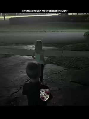 Two children are shown in a street scene at nighttime. A boy in the foreground faces away from the camera with a Porsche crest on his back. A girl stands further away, facing away with her arms outstretched. The scene is dark and the lighting suggests it is nighttime.