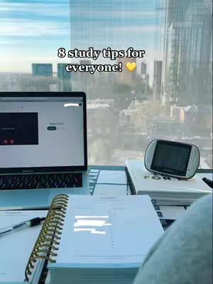 The image shows a desk setup with a laptop displaying a screen and a notebook with a gold spiral binding. The scene is set in front of a large window with a city skyline visible outside. Text overlaid on the image reads: "8 study tips for everyone!" with a yellow heart emoji.