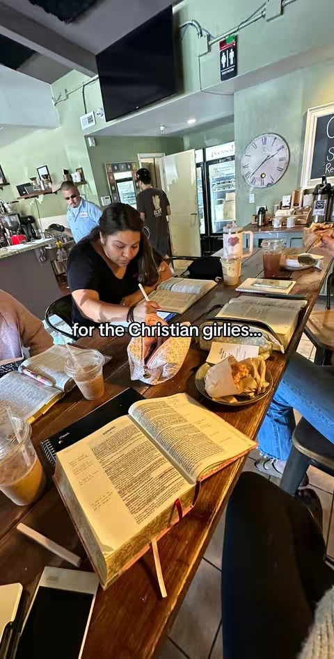 A medium shot shows a table in a cafe setting with multiple people. Books and notebooks are spread across the wooden table. A woman is writing with a stylus on a tablet. A coffee cup is partially visible, and there are plates with food. The background shows a wall painted in pastel shades and a clock. The lighting is natural, suggesting daytime.