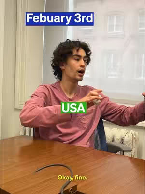 A man in a pink shirt sits at a wooden table, gesturing with his hands. Text overlays indicate the date and the USA. The image conveys an expression of mild frustration or resignation.