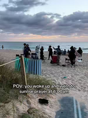 A photograph taken at dawn shows a beach scene with multiple figures gathered. The sky is a blend of pastel colors, and the ocean stretches to the horizon. People are set up in chairs on the beach and some are raising their hands. The foreground shows sand and some greenery.