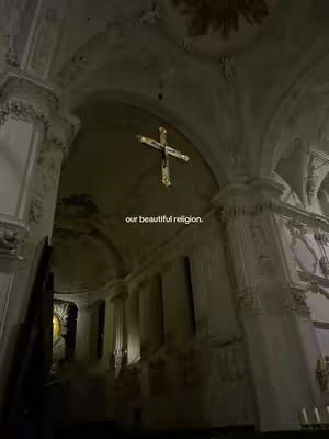 An interior shot of a cathedral or church. The focus is on a cross illuminated at the highest point of the arch. The lighting is soft and dramatic emphasizing the ornate architectural details. The image uses a muted color palette with a slight vignette effect.