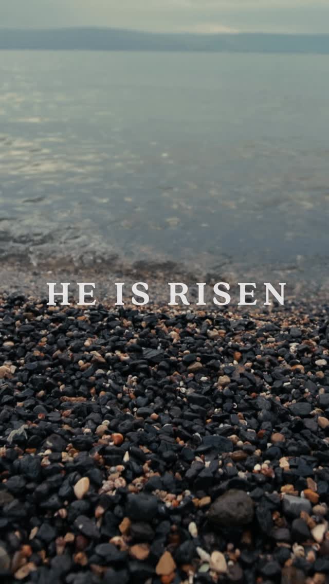 A serene image of a shoreline with water and rocks. In the foreground, small, dark rocks cover the lower portion of the frame, transitioning to the water of the Sea of Galilee in the mid-ground. The sky is overcast, with pale colors adding a calming effect. The text overlay, 'HE IS RISEN,' is centered and in a clean, white font.