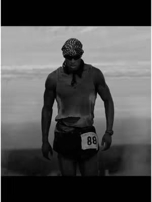 A black and white image of a muscular man standing outdoors. He is wearing a dark bandana, sunglasses, a tank top, and shorts with a race bib. The background is blurred, suggesting open space.