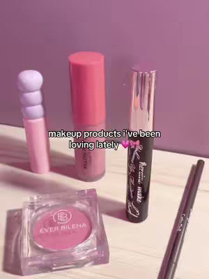 The cover image showcases an assortment of makeup products arranged on a light-colored surface against a blurred purple background. The products include a pink lip gloss, a mascara tube, an eye pencil, and a blush compact. Text overlayed on the image reads "makeup products i've been loving lately" and a pink heart emoji.