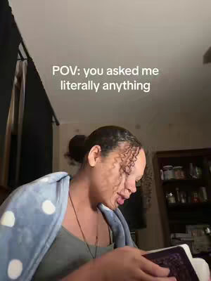 A woman with dark hair, styled in a bun, is looking down at a book or document in her hands. She is framed from the chest up, seated indoors with a blanket over her shoulders. A text overlay at the top of the image reads 'POV: you asked me literally anything'.