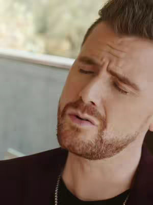 A close-up of a man's face, slightly angled, with closed eyes and a pensive expression. He has a beard and short, dark hair. He is wearing a dark maroon blazer over a black shirt, and appears to be indoors.