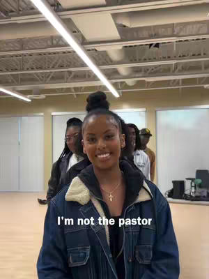 A woman smiles at the camera, with a line of people behind her. Text overlayed reads "I'm not the pastor". The woman is wearing a light colored jacket over a dark shirt and a cross necklace. Behind her are a few other figures, also dressed, but somewhat faded into the background.