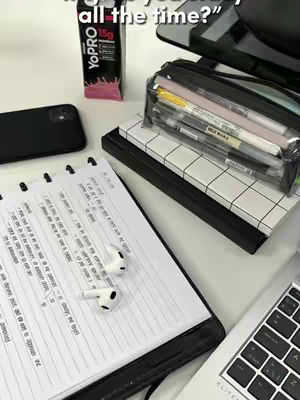 The image shows a study scene with a notepad, pens, headphones, and a laptop. The notebook is open with handwritten notes and headphones next to it. A laptop is partially visible, along with a phone, a drink container and stationary.