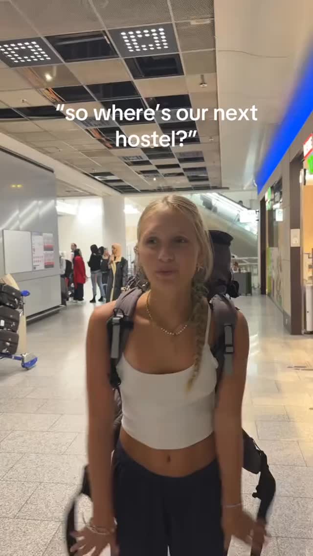 A young woman with blonde hair, wearing a white top and dark pants, stands in a well-lit airport hallway. She is carrying a backpack and looking towards the camera. The background shows other travelers, luggage, and signs.