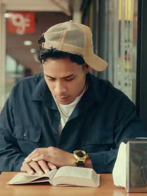 A young man is seated at a table, looking down at an open book. He wears a casual outfit including a cap and a watch. The background is softly blurred, suggesting an indoor setting.
