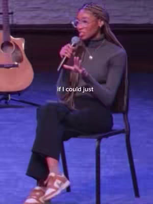 A woman with long braids wearing glasses and a turtleneck sits on a chair, holding a microphone. She is gesturing with her hands as if speaking in front of an audience. A guitar is visible in the background, and some text 'if I could just' is displayed over her image