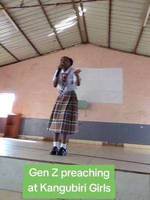 A young person, likely a student, is standing on a stage giving a speech. They are holding a microphone and gesturing with one hand. The background is a simple tan-colored wall, with a white rectangle present. Below the stage, a green banner highlights text.