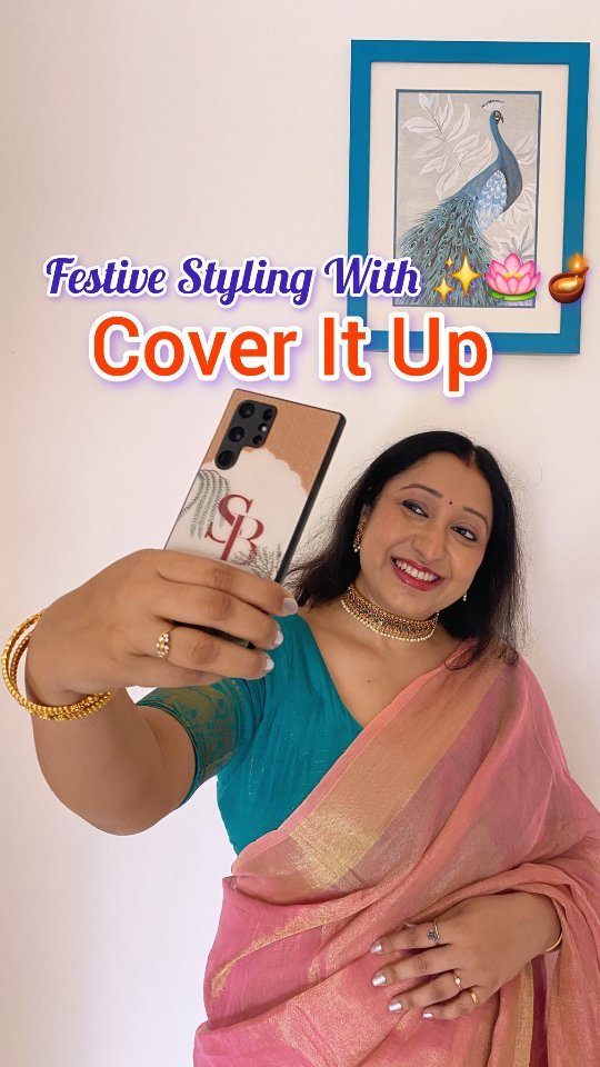 A woman is holding a phone showing its customized cover with her initials looking at the camera with a smile. The cover is a shade of brown with a few drawings of leaves in a red color and her initials, SB. She is wearing a turquoise shirt and a pink saree. In the background, there is a framed picture of a peacock. The hook text is overlaid on the top.
