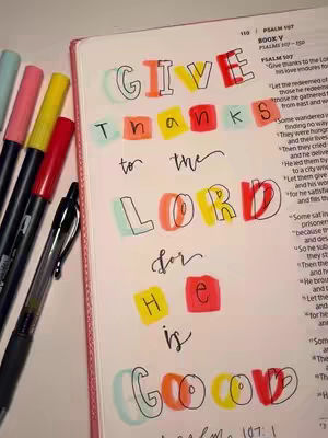 An open Bible with text highlighted in colorful pens, with the words "Give Thanks to the Lord for He is Good" written in a handwritten style on the pages.