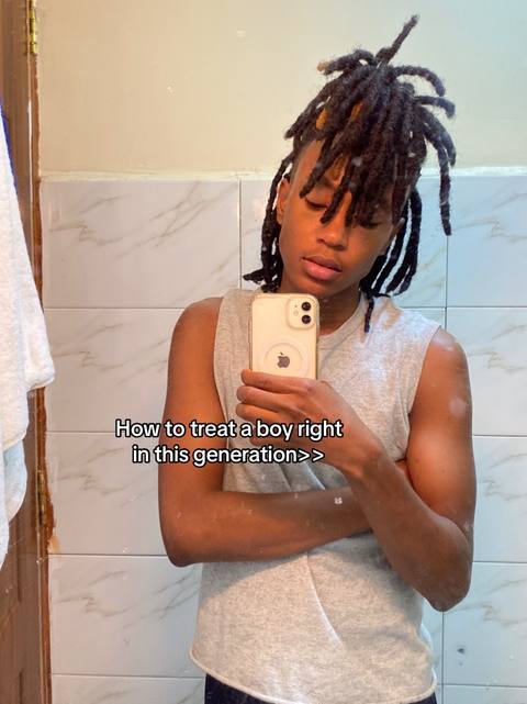 The selfie-style photo features a person with dark hair and dreadlocks holding a phone in front of a bathroom mirror. The background includes a light-colored wall with a patterned tile border and a white towel hanging on a bar. The person is wearing a light gray sleeveless shirt.