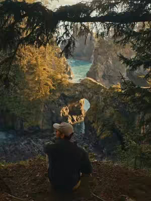 A scenic landscape featuring a person gazing out at a natural archway over a body of water. The person is wearing a baseball cap and dark clothing, seen from the back. The foreground is earthy with dark soil, while the background reveals ocean and rock formations. The lighting appears to be natural, with a mix of shadows and sunlight.