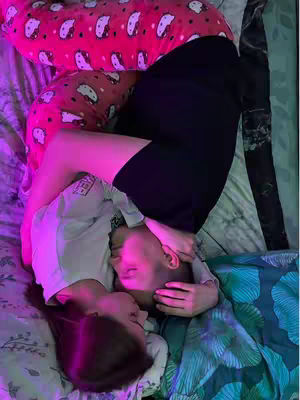 Two people, a male and a female, are embracing while laying on bed covered by a pink Hello Kitty themed pillow and a teal floral print blanket. They are lying on top of each other, with their faces close. The lighting in the room suggests a purple or pink hue, creating a soft, intimate atmosphere. The composition of the image is centered around the couple, highlighting their connection.
