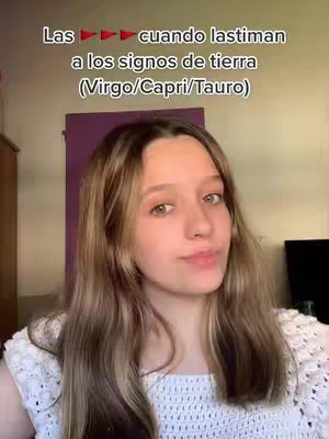 A young woman with long, light-brown hair, wearing a white crocheted top, is centered in the frame. The background includes a purple panel and a glimpse of a wooden cabinet. Text overlays at the top indicate the video's topic, discussing earth zodiac signs.