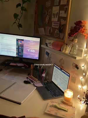 An interior shot of a study space with a monitor, a laptop, books, and other stationery. The background feels warm and inviting, with soft lighting from fairy lights and a candle.