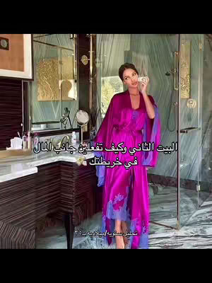 A woman in a luxurious bathroom wearing a long, flowing pink robe with blue accents. She talks close to a mirror next to a counter and a shower partition.