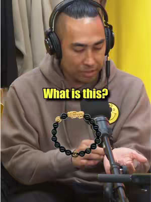 A man wearing headphones is looking down with an introspective expression. Text that reads "What is this?" in a yellow text box hovers above the man. In front of the man is a microphone and a bracelet.