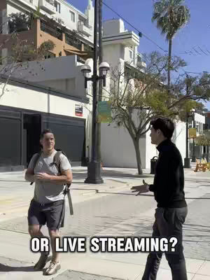 Two men are on a sidewalk, facing each other with a city street and buildings in the background.  One man is casually dressed while the other is wearing a long sleeve black shirt and pants. Text is overlaid on the bottom of the image.