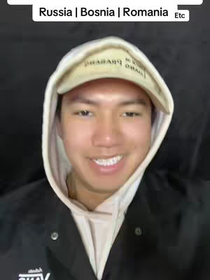 A young man with fair skin is shown from the chest up, centered in the frame. He wears a white hoodie under a black jacket, with a beige cap that has words printed on its front. The man is smiling with his teeth showing. Above him, there is a text box with the labels: Russia | Bosnia | Romania | Etc.