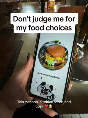 A person is holding a smartphone displaying an app interface. The app shows a photo of a burger in a bowl, with text overlays that read, "Don't judge me for my food choices." The phone screen also displays a cartoon raccoon and text below the burger image, with details about nutrition and tips.