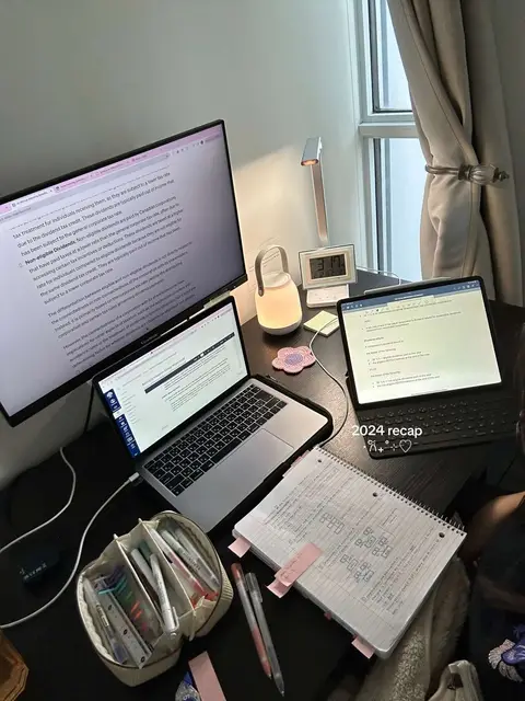 A top-down shot of a desk setup with three screens, a laptop, a pen case, notebooks, and a digital clock. The image has a warm, cozy aesthetic, with soft lighting and neutral colors.