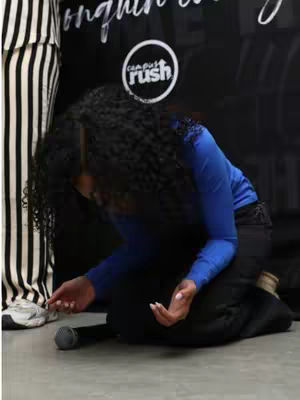 A person kneels down with their head bowed, appearing to be in a position of worship. The background features a black surface, a partial view of what looks like a sign or backdrop, and a partial view of someone wearing a black and white striped garment. The lighting appears to be even.