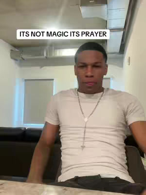 A man in a white t-shirt and silver chain sits in front of a dark couch. The background shows an industrial ceiling and partially visible room. The text overlay says "ITS NOT MAGIC ITS PRAYER".