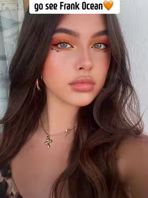 A close-up shot of a woman with elaborate, colorful eye makeup and long, dark brown hair. She has pale skin, full lips, and a cross-shaped pendant necklace.