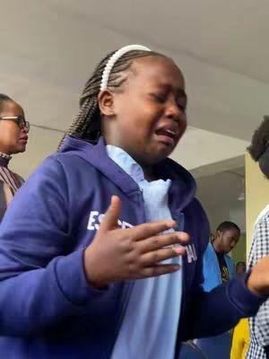 A young girl is crying, with tears streaming down her face and with an open mouth. She appears to be in some sort of public space with other people around. She is wearing a blue hoodie over a white shirt, a white headband and braided hair. The background is blurred and suggests an indoor setting.
