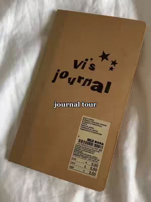A brown notebook with the title 'Vi's journal' in black text. A hook 'journal tour' is displayed underneath the title. The notebook has a sticker on it and it is set on a white surface.