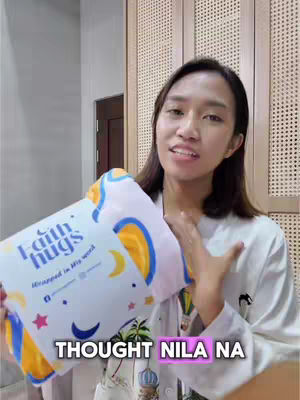 A woman is holding a folded white and blue blanket with the text 'faith hugs' printed on it. She is smiling at the camera, with text overlayed at the bottom in pink. The background is a home interior with wooden panels and a door.