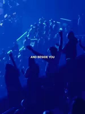 A vertical video taken at a concert setting. The image is dominated by a blue color palette, and many people are visible, their arms raised. The shot is from a low angle, adding to the elevated atmosphere and a sense of community and participation.