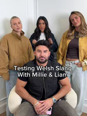 A group of four people, three women and one man, standing in a well-lit room. The man is in the foreground seated, while the women are behind him. Text in a contrasting white box at the bottom of the cover image reads, "Testing Welsh Slang With Millie & Liam".