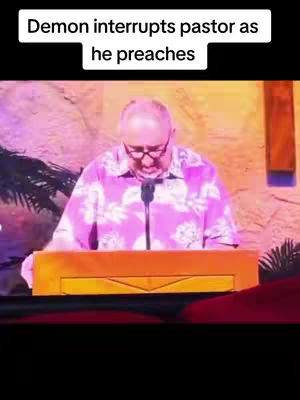 A man in a pink floral shirt stands behind a wooden podium, speaking into a microphone. The background appears to be part of a stage. Text overlay at the top says, "Demon interrupts pastor as he preaches".