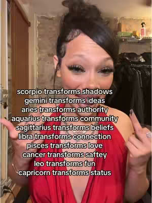 A woman with a high bun hairstyle and heavy eye makeup is centered, pointing with her left hand while her right hand makes a peace sign. The background includes glimpses of a room with what seems to be shelves and a door.