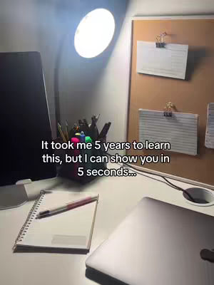 An overhead shot captures a well-lit desk setup. A bright lamp illuminates the scene from above, casting light on a computer screen, a pen holder with pens and markers, a notebook with a pen resting on it, and a laptop. A corkboard with index cards clipped to it is visible, as well as additional desk items, suggesting a study or work environment.
