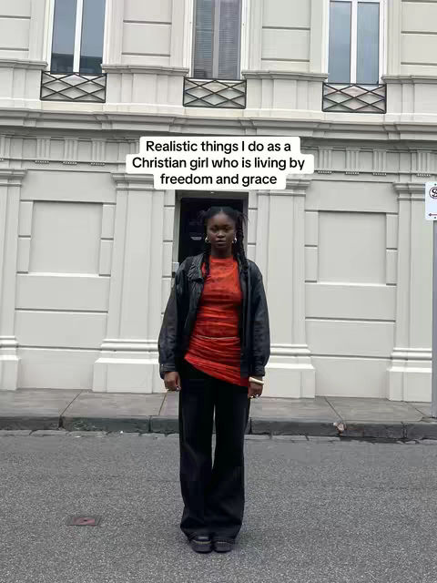 A full shot against an urban building backdrop. A woman of color is the focal point, centered and standing still on a gray paved surface. She wears a black leather jacket over a red patterned top and black pants. The background building is a light color, with evenly spaced windows and decorative elements, suggesting a European architectural style. The overall composition gives a sense of order and simplicity, suitable for inspirational content.