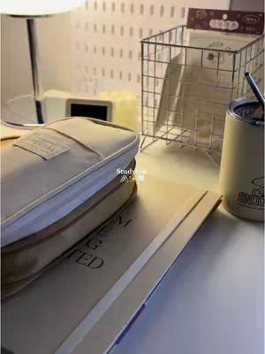 A well-lit desk setup with neutral tones. A beige pencil case sits on top of a closed book, and there is a pen next to an open notebook. A snoopy cup is next to other items on the desk.