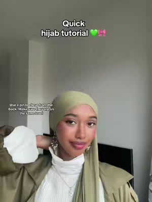 A woman wearing a hijab, facing forward with a slight smile. Text overlays the image instructing the viewer on how to wear a hijab. The woman is wearing a green hijab and top with a white top inside, and jewelry. The background is a plain wall with a dark object and a bright light source.