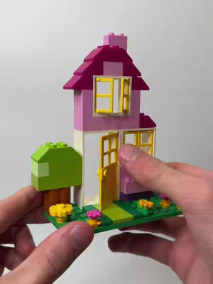 A person's hands hold a small house constructed from colorful Lego blocks. The house is primarily pink, with a red roof and yellow windows and doors. To the left, a green block represents a tree. The background is a plain white surface.