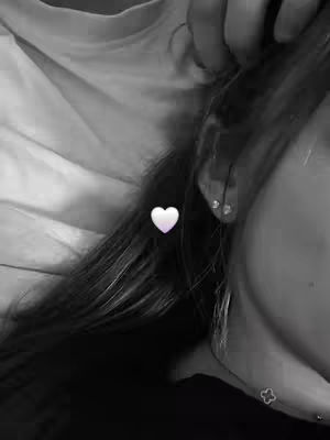 A close-up, black and white image. The focus is on the side of a person's face, from the ear to the bottom of the cheek. Long, dark hair flows across the image. A white heart sticker is placed between the hair and the ear.