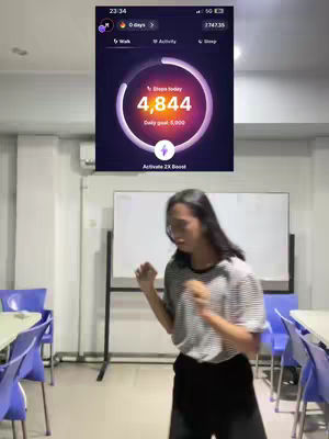A young woman stands in the middle of a brightly lit room, likely an office space or classroom. She is smiling, and her arms are bent as though she is lightly dancing. On the wall above her is a screen showing the Sweatcoin app, partially obscuring the top of the frame. There is an active step counter on the screen showing 4,844 steps.
