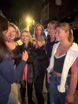 A group of six young women are standing closely together on a dimly lit street at night. The woman in the foreground on the left is holding a microphone and appears to be interviewing the other women. The women are smiling and laughing, suggesting a casual and fun atmosphere. Behind them, there's a street with buildings and a street light adding to the urban setting.