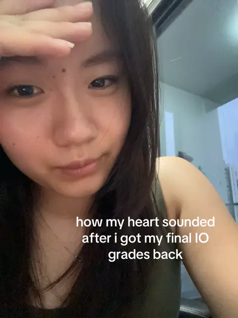 A selfie-style cover image features a young Asian woman with dark hair, looking at the camera. Her right hand is raised to her forehead, and a soft, slightly desaturated color palette dominates, with the background containing elements of a window or a light source. Text overlayed on the bottom of the image reads 'how my heart sounded after I got my final IO grades back,'