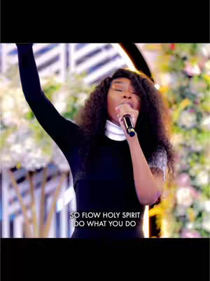 A woman with long, curly hair is singing into a microphone with her arm raised. She is wearing a black outfit with a white collar. Text overlayed at the bottom says: SO FLOW HOLY SPIRIT DO WHAT YOU DO.
